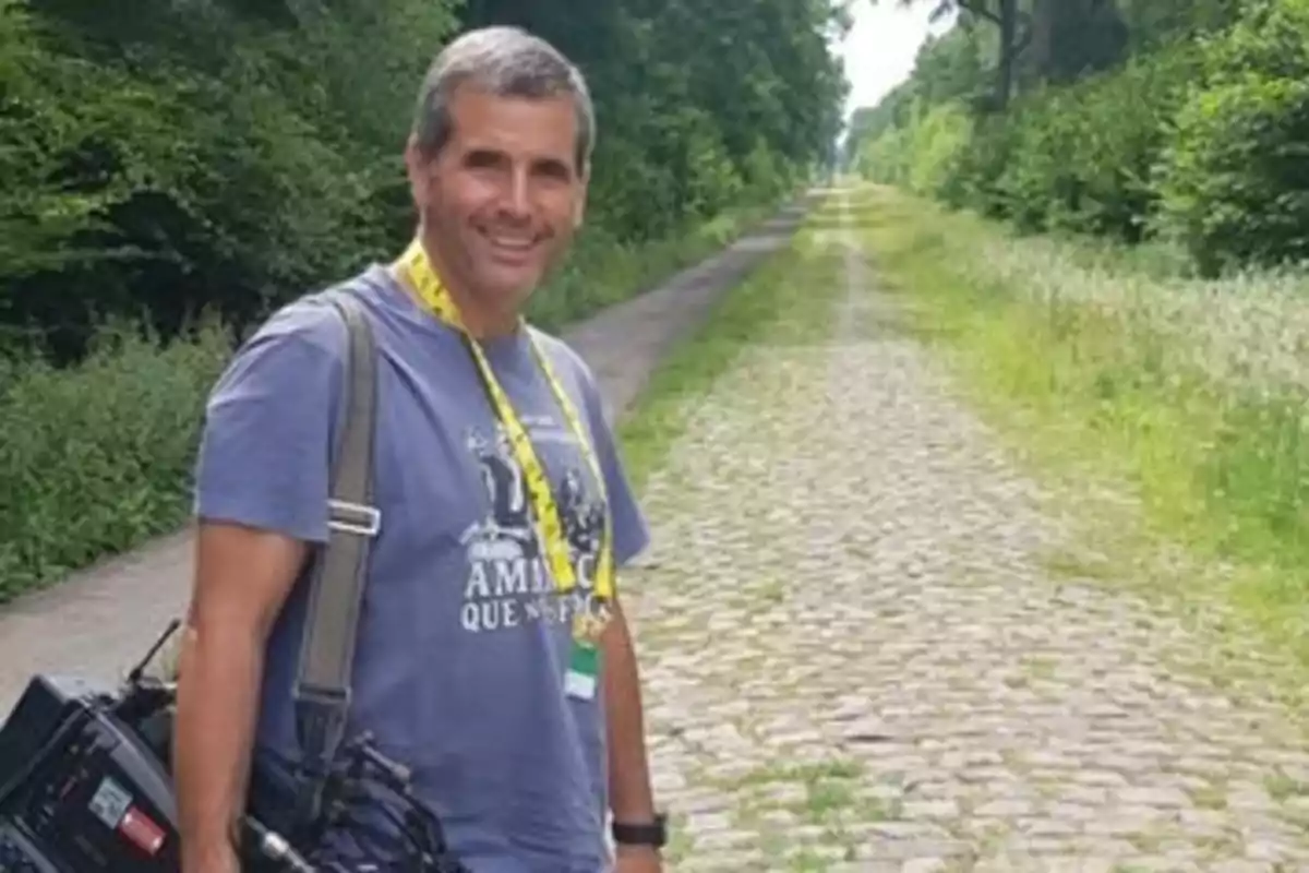 Hombre sonriente con camiseta azul y cámara al hombro parado en un camino de adoquines rodeado de vegetación Hombre sonriente con camiseta azul y cámara al hombro parado en un camino de adoquines rodeado de vegetación