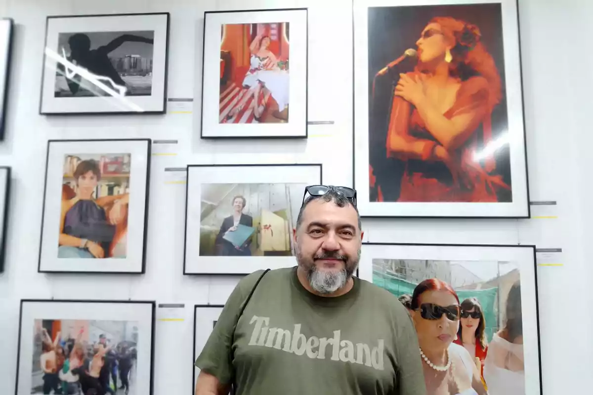 Hombre de barba y camiseta verde con la palabra Timberland posando frente a una pared llena de fotografías enmarcadas en una galería de arte Hombre de barba y camiseta verde con la palabra Timberland posando frente a una pared llena de fotografías enmarcadas en una galería de arte