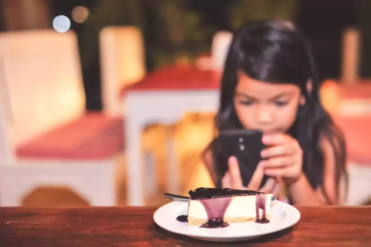 Una niña enfocada en su teléfono móvil mientras una rebanada de pastel de queso con salsa de arándanos está en primer plano sobre una mesa.