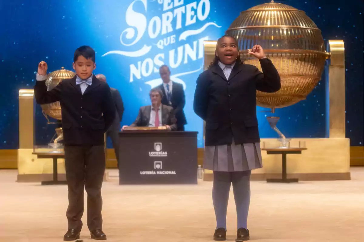 Dos niños en uniforme escolar participan en un sorteo de lotería, sosteniendo bolas numeradas frente a un gran bombo dorado, con un fondo azul que dice 