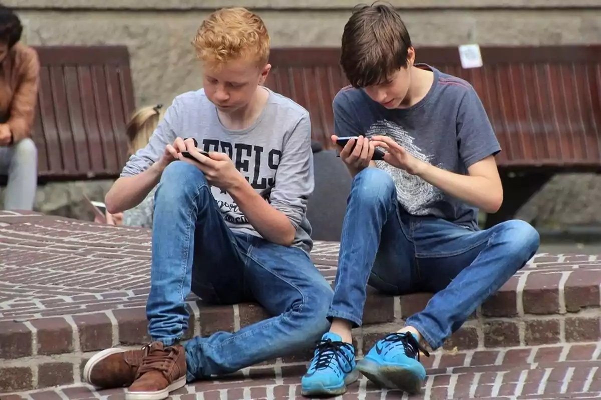 Dos adolescentes sentados en una escalera de ladrillos usando sus teléfonos móviles.