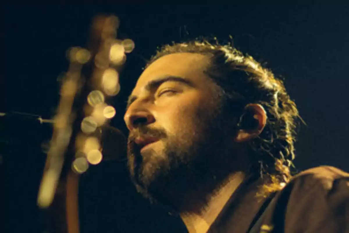 Un hombre con barba y el cabello recogido canta con los ojos cerrados mientras toca la guitarra en un escenario iluminado.