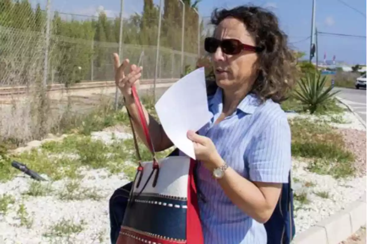 Una mujer con gafas de sol sostiene un papel y un bolso mientras está de pie al aire libre cerca de una carretera y una cerca.