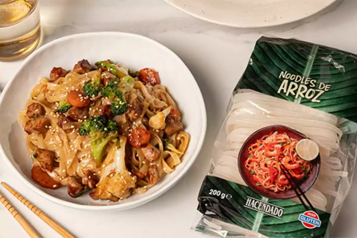 Un plato de noodles de arroz con verduras y carne junto a un paquete de noodles de arroz sobre una mesa. Un plato de noodles de arroz con verduras y carne junto a un paquete de noodles de arroz sobre una mesa.