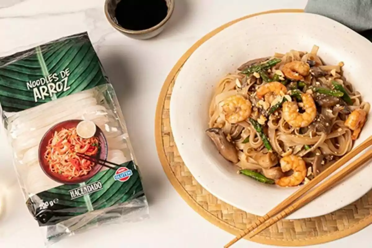 Un plato de noodles de arroz con camarones, espárragos y champiñones junto a un paquete de noodles de arroz y un tazón de salsa sobre una mesa. Un plato de noodles de arroz con camarones, espárragos y champiñones junto a un paquete de noodles de arroz y un tazón de salsa sobre una mesa.