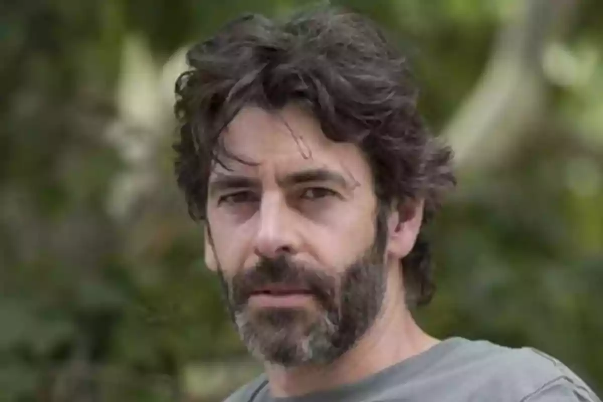 Hombre con barba y cabello oscuro mirando a la cámara con un fondo de naturaleza.