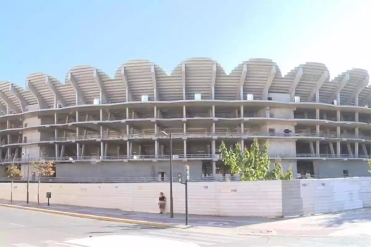 Edificio en construcción con estructura de concreto y techos curvados, rodeado por una valla blanca y ubicado junto a una calle.