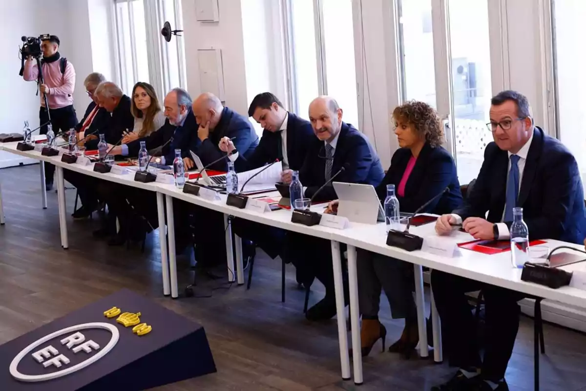 Un grupo de personas sentadas en una mesa de conferencias con documentos y botellas de agua, mientras un fotógrafo captura el momento.