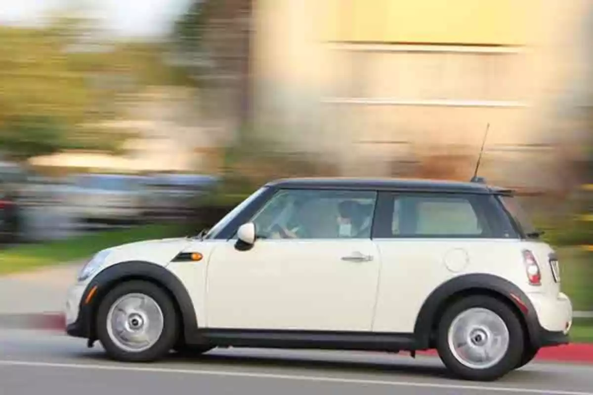 Un coche compacto blanco circula rápidamente por una calle con el fondo desenfocado.