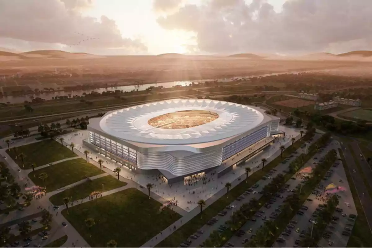 Vista aérea de un estadio moderno al atardecer, rodeado de áreas verdes y estacionamientos, con colinas y un río en el fondo.