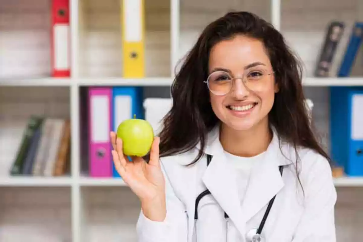 Mujer con bata de laboratorio y estetoscopio sosteniendo una manzana verde, sonriendo frente a una estantería con carpetas de colores.