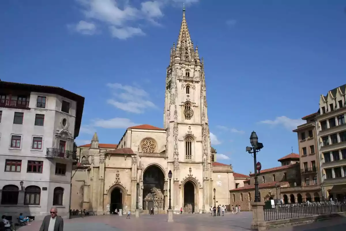 Una catedral gótica con una torre alta y detallada se alza en una plaza rodeada de edificios históricos bajo un cielo azul.