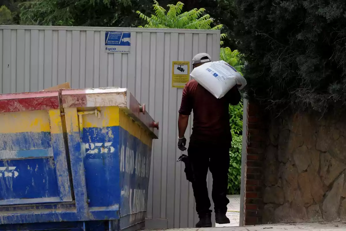 Un hombre cargando un saco en el hombro entra por una puerta metálica junto a un contenedor de basura.