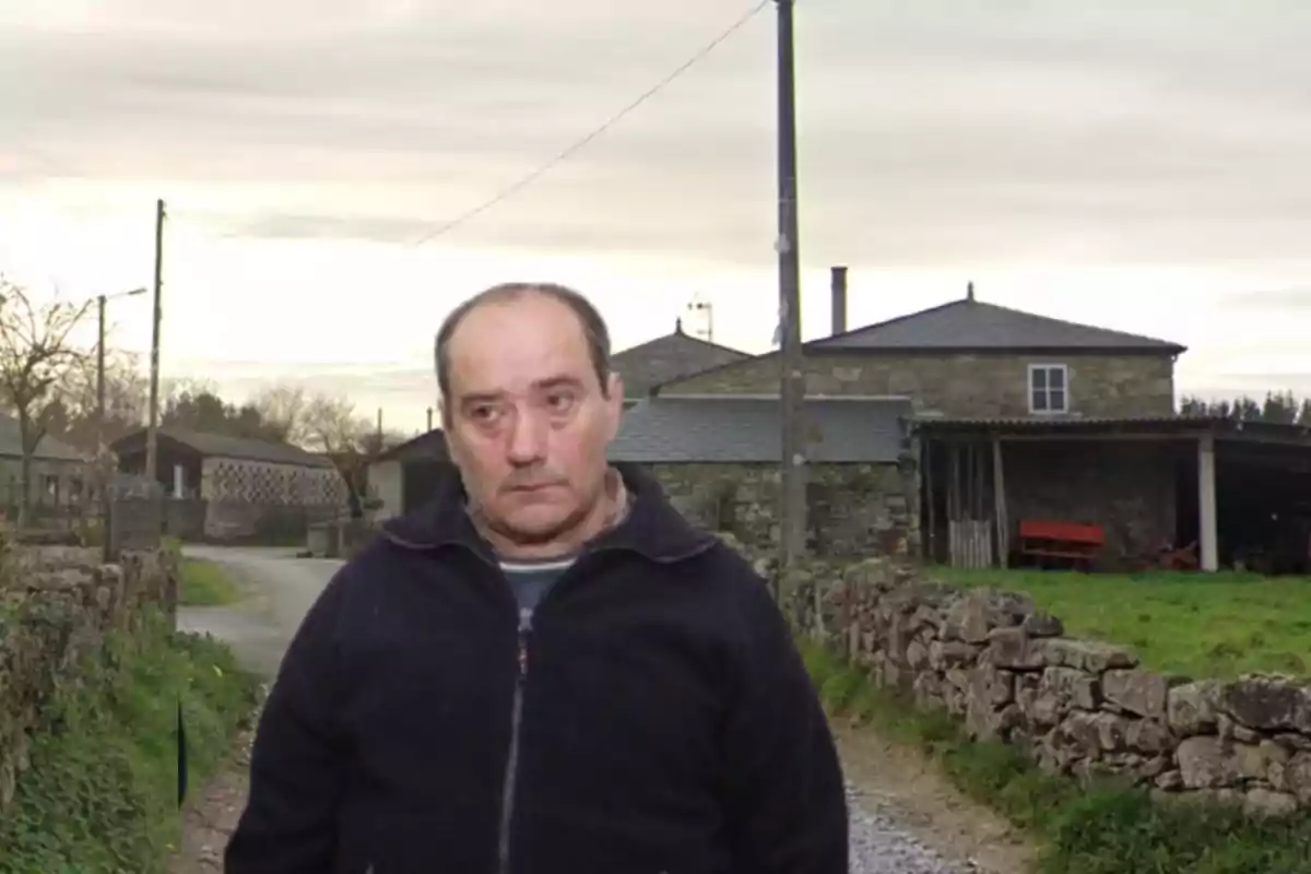 Un hombre de pie en un camino rural con casas de piedra al fondo.