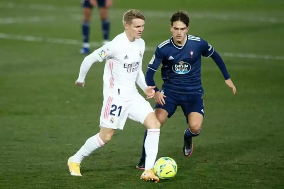 Dos futbolistas compiten por el balón en un partido de fútbol, uno viste de blanco y el otro de azul. Dos futbolistas compiten por el balón en un partido de fútbol, uno viste de blanco y el otro de azul.