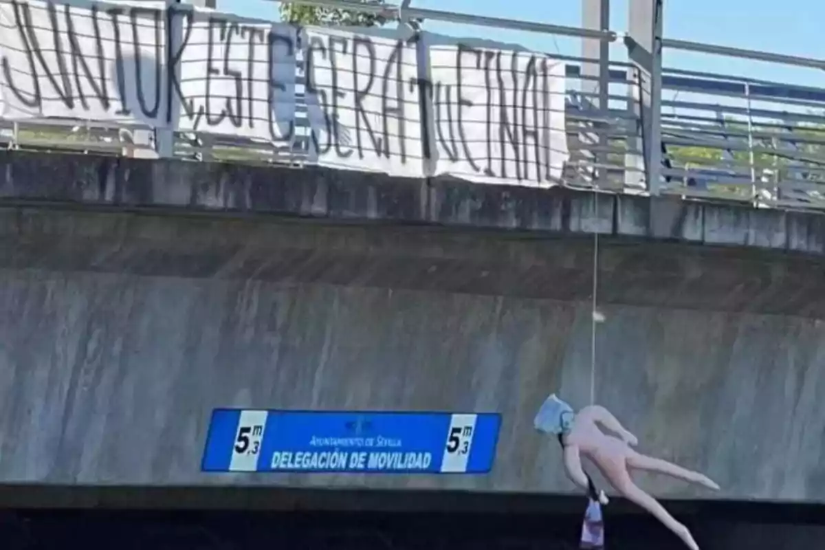 Un puente con una pancarta colgada que tiene un mensaje escrito en letras grandes y una figura inflable suspendida debajo del puente. Un puente con una pancarta colgada que tiene un mensaje escrito en letras grandes y una figura inflable suspendida debajo del puente.
