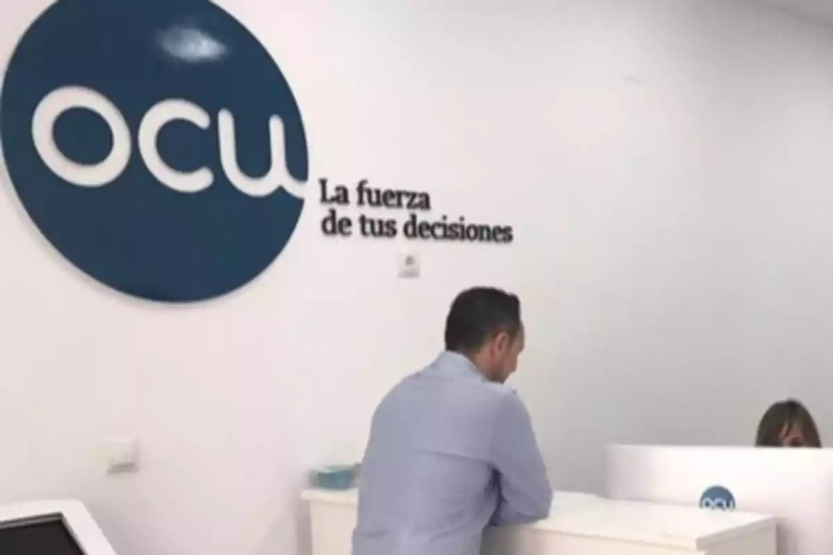 Un hombre de pie frente a un mostrador en una oficina con el logotipo de OCU y el lema "La fuerza de tus decisiones" en la pared.