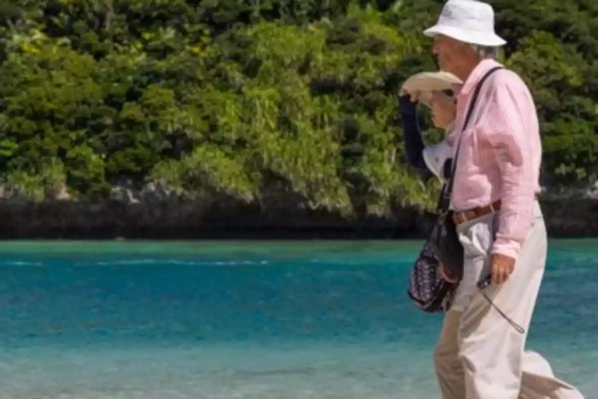 Dos personas mayores caminando en la playa con sombreros y ropa ligera, con un fondo de vegetación y mar azul.