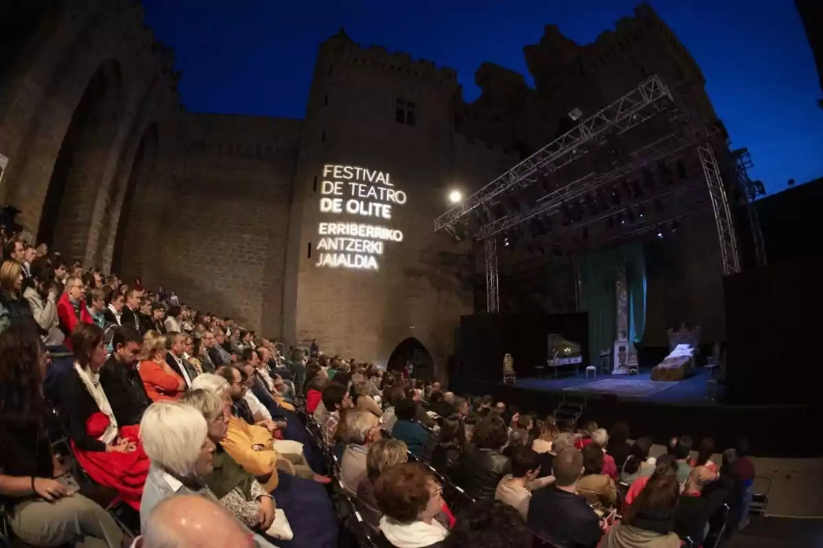 Una audiencia observa una obra de teatro en un escenario al aire libre durante el Festival de Teatro de Olite, con una proyección en la pared que indica el nombre del evento en español y euskera.