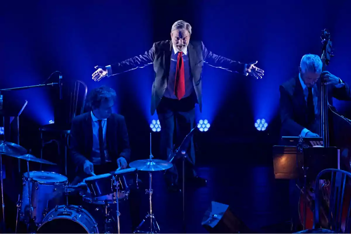 Un hombre con traje y corbata roja está de pie con los brazos extendidos en un escenario iluminado de azul, acompañado por un baterista y un contrabajista.