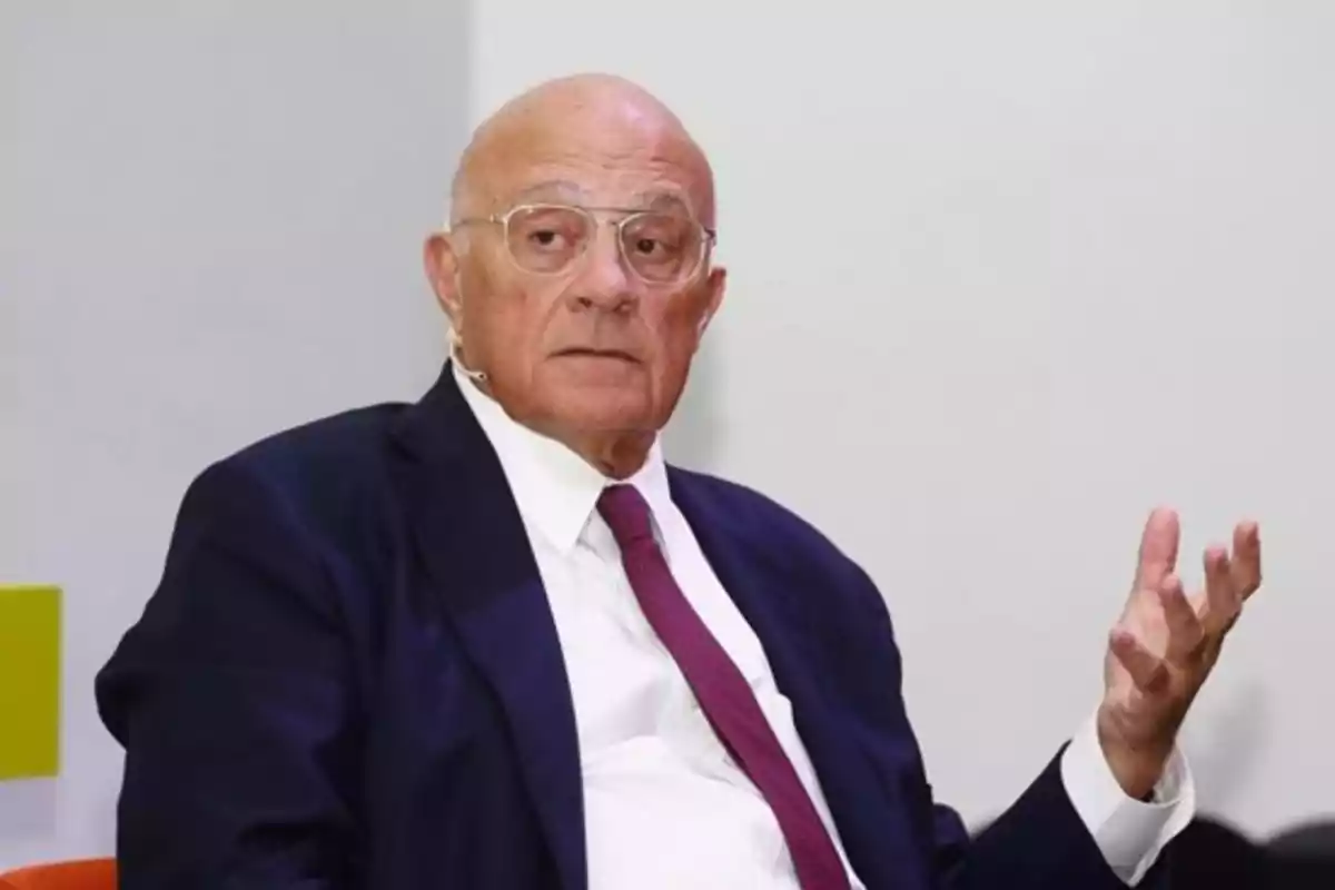 Un hombre mayor con gafas y traje oscuro gesticula mientras está sentado. Un hombre mayor con gafas y traje oscuro gesticula mientras está sentado.