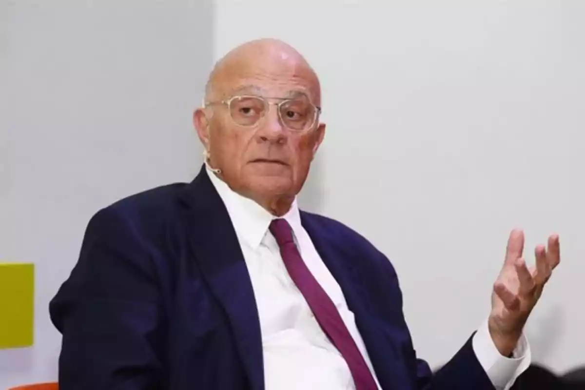 Hombre mayor calvo con gafas y traje oscuro sentado y gesticulando con la mano Hombre mayor calvo con gafas y traje oscuro sentado y gesticulando con la mano