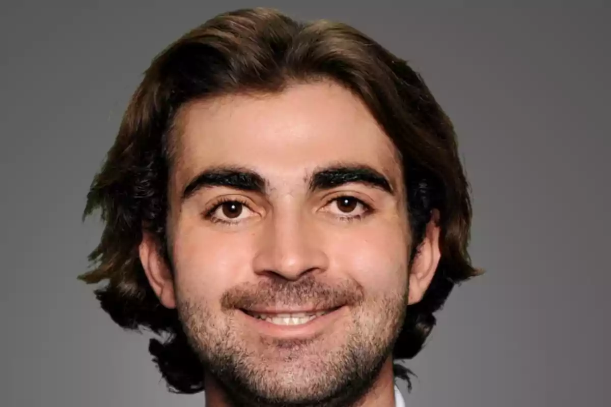 Hombre joven de cabello castaño y barba ligera sonriendo frente a un fondo gris Hombre joven de cabello castaño y barba ligera sonriendo frente a un fondo gris