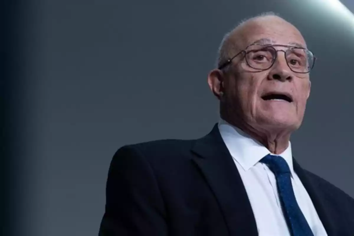 Hombre mayor con gafas y traje oscuro hablando frente a un fondo gris Hombre mayor con gafas y traje oscuro hablando frente a un fondo gris