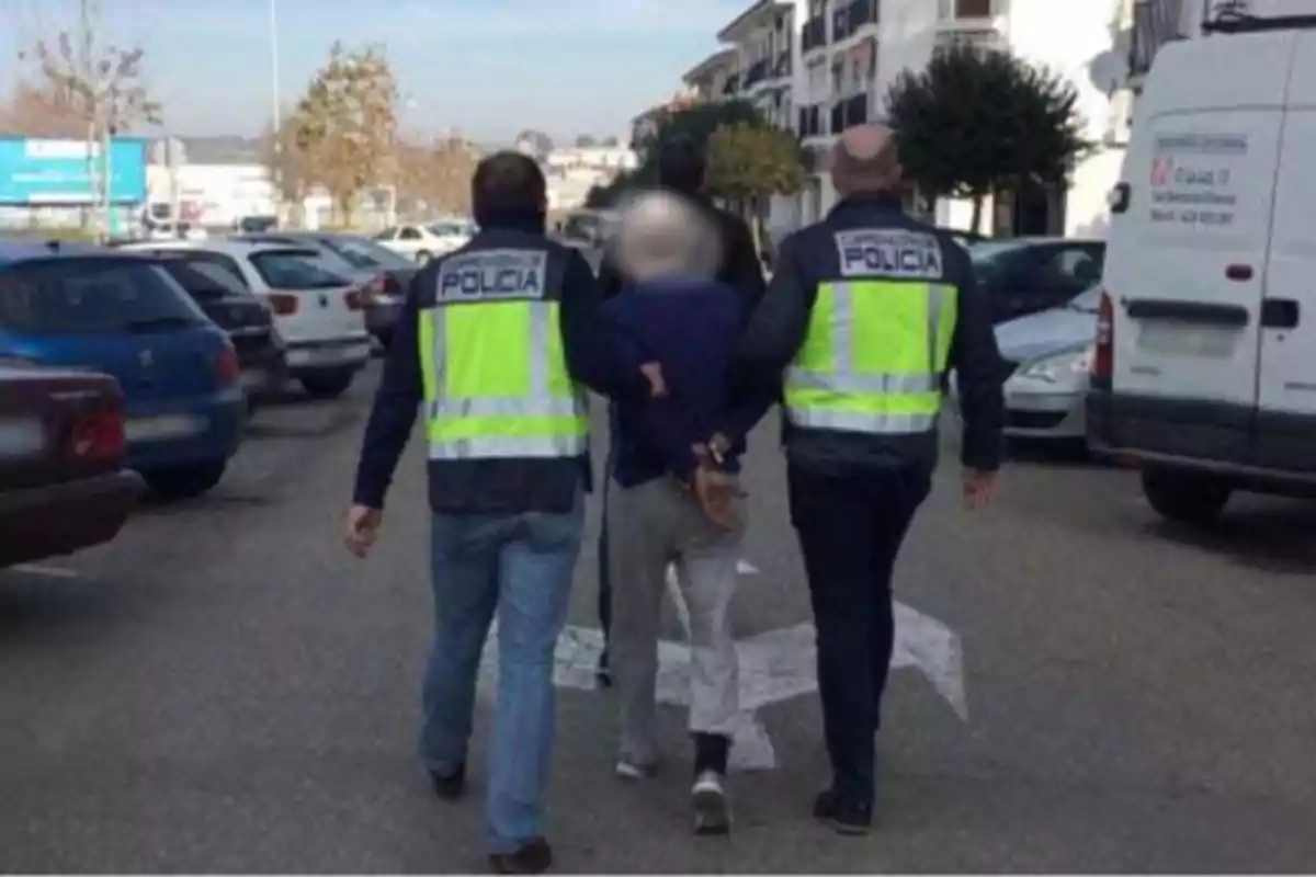 Dos policías escoltan a una persona esposada por una calle con varios coches estacionados. Dos policías escoltan a una persona esposada por una calle con varios coches estacionados.