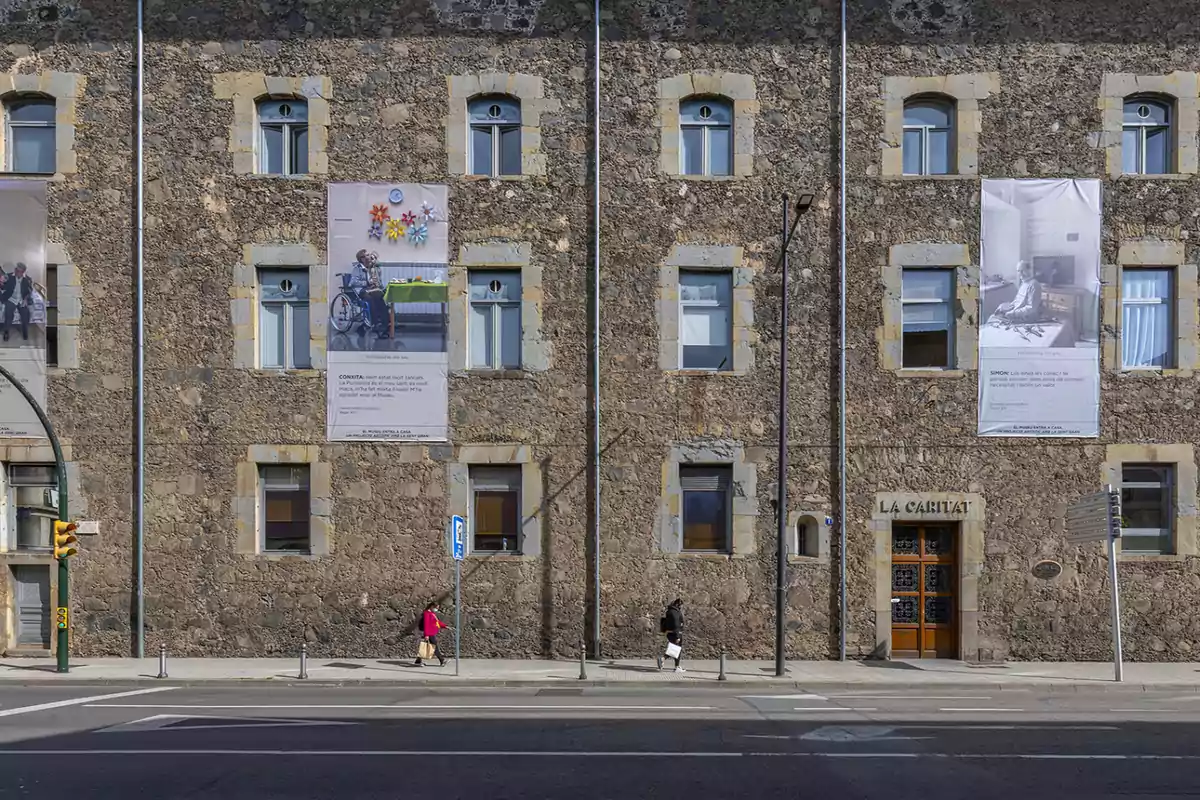 Fachada de un edificio de piedra con varias ventanas y carteles grandes colgados en la pared, dos personas caminando por la acera.