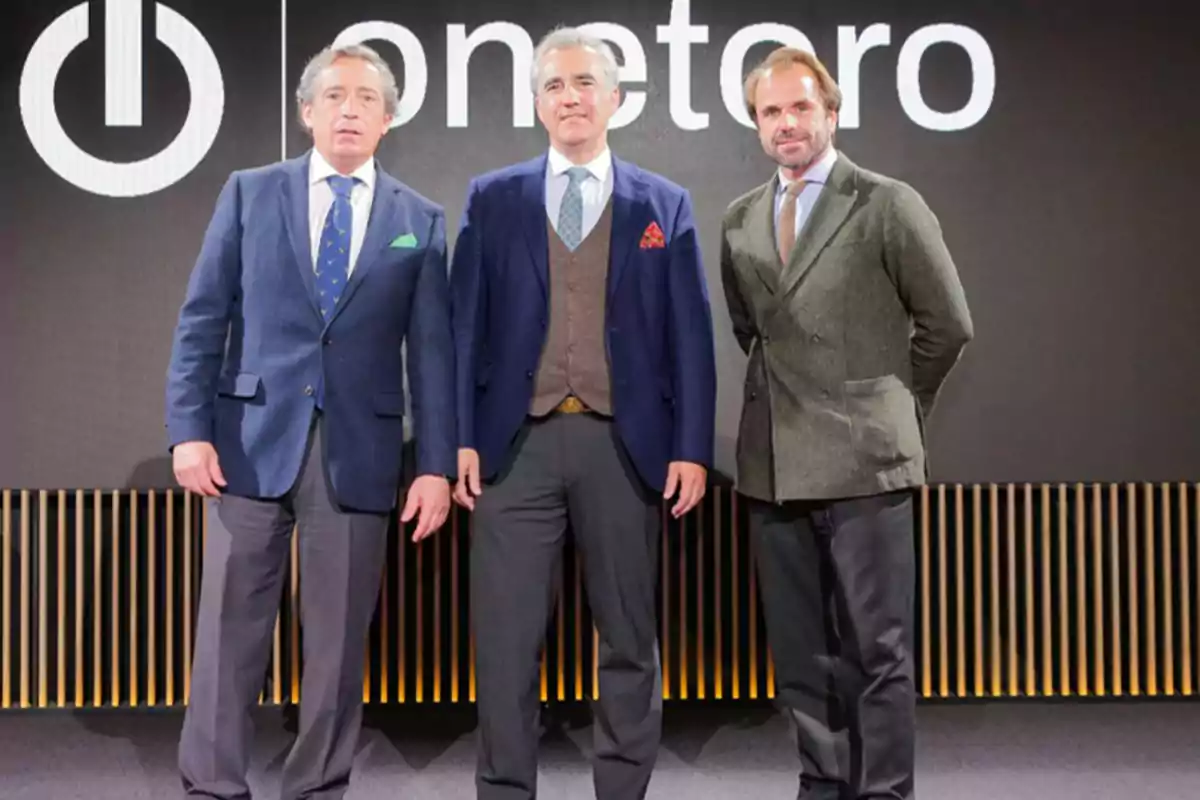 Tres hombres de pie, vestidos con trajes formales, posan frente a un fondo con el logotipo de "onetoro".