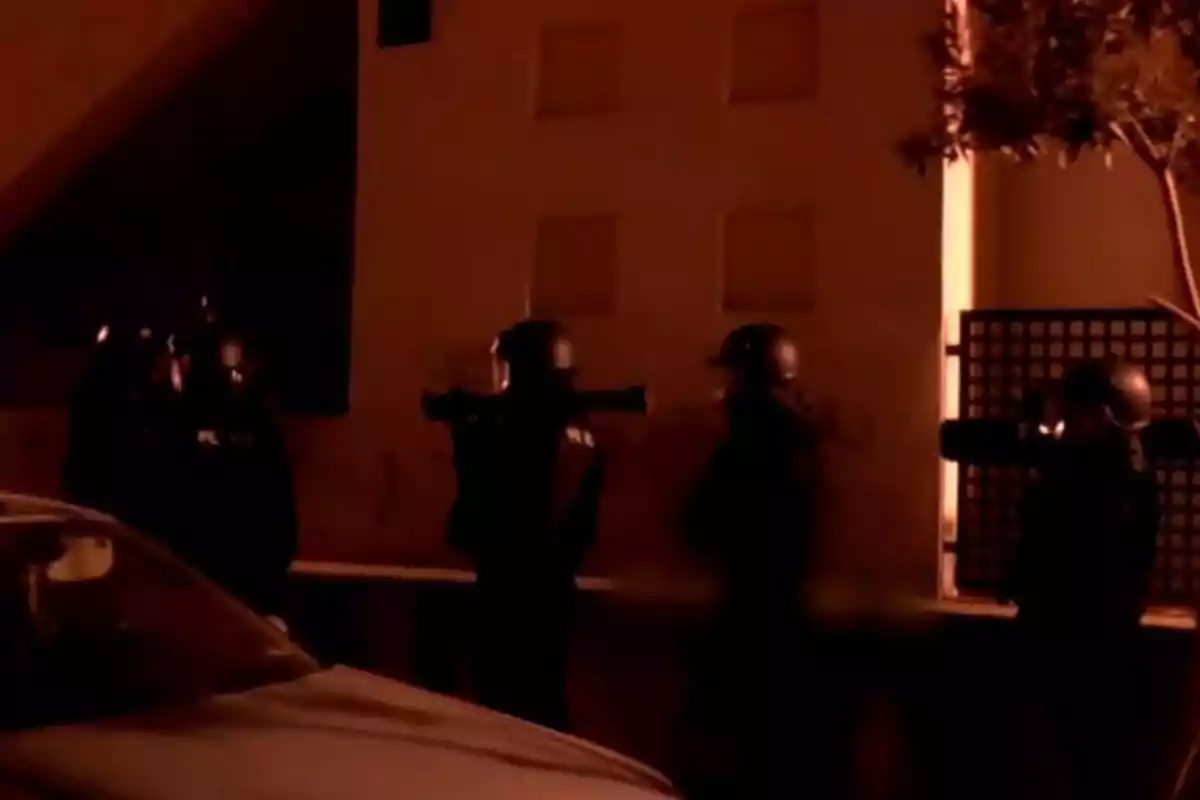 Un grupo de personas con cascos y equipo táctico se encuentra de pie junto a un edificio en una escena nocturna iluminada con luz anaranjada.