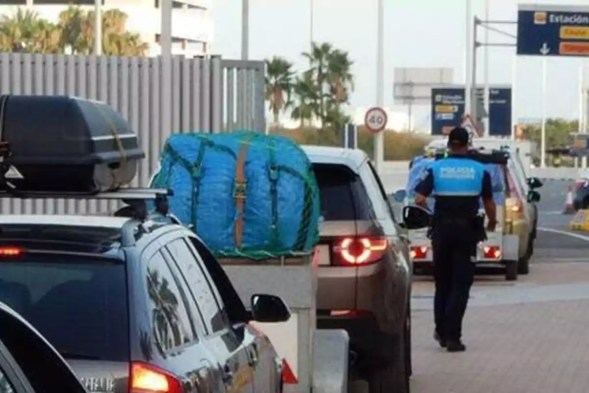 Un agente de policía de tráfico revisa vehículos en una carretera, donde se observan coches con equipaje en el techo y remolques.