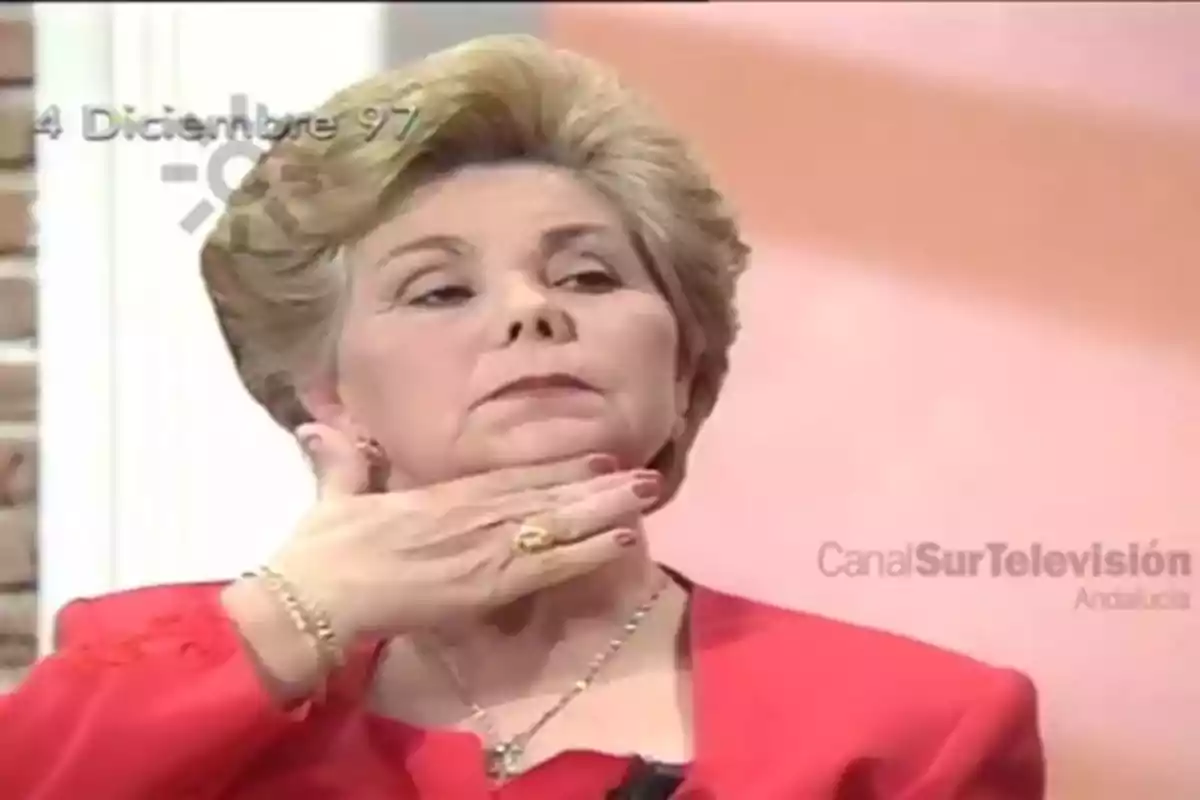 Una mujer con cabello rubio y un traje rojo se toca el cuello mientras está en un programa de televisión.