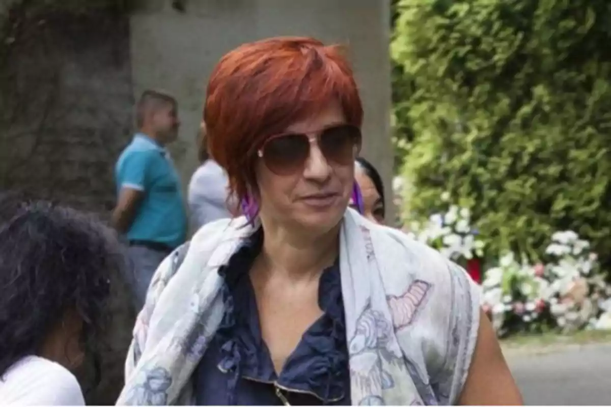 Mujer con cabello corto y rojo, usando gafas de sol y una bufanda clara, en un entorno al aire libre con personas y vegetación de fondo.