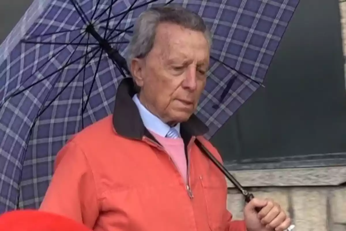 Ortega Cano con chaqueta naranja sostiene un paraguas a cuadros. Ortega Cano con chaqueta naranja sostiene un paraguas a cuadros.