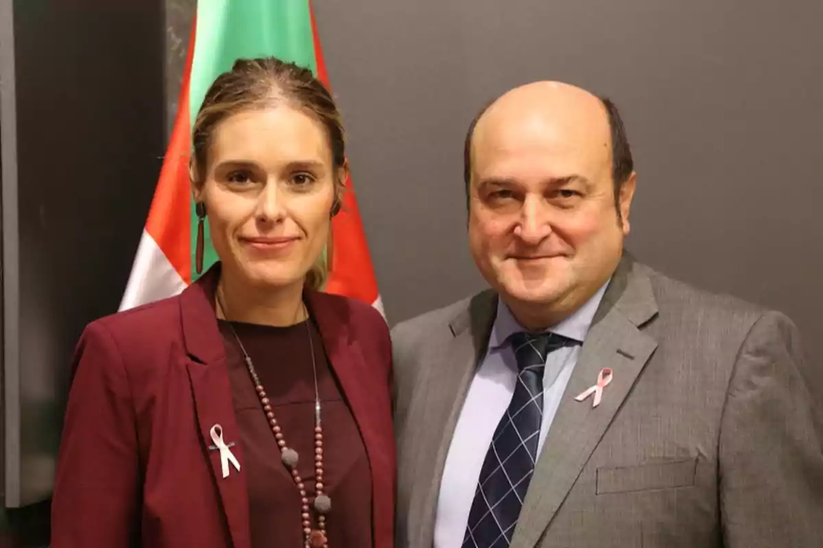 Dos personas posan juntas frente a una bandera, ambas llevan lazos rosas en sus prendas.