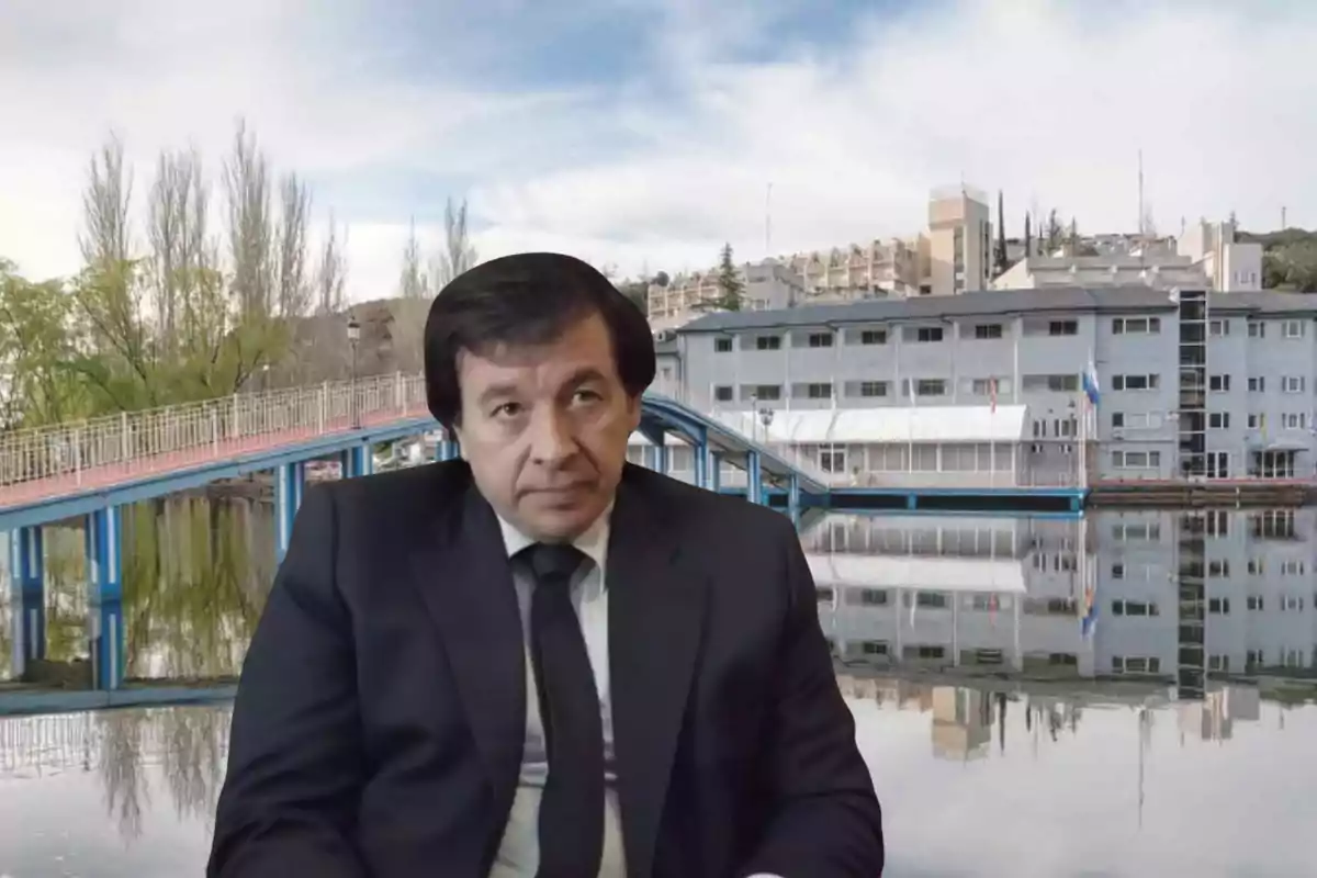 Un hombre con traje oscuro está sentado frente a un fondo de un puente azul y un edificio junto a un lago.