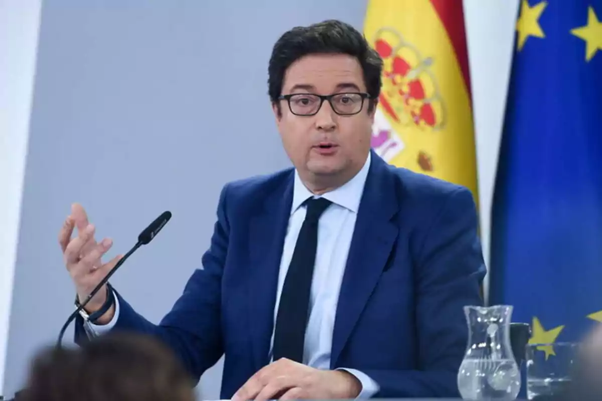 Hombre con traje azul hablando en una conferencia de prensa con las banderas de España y la Unión Europea de fondo.