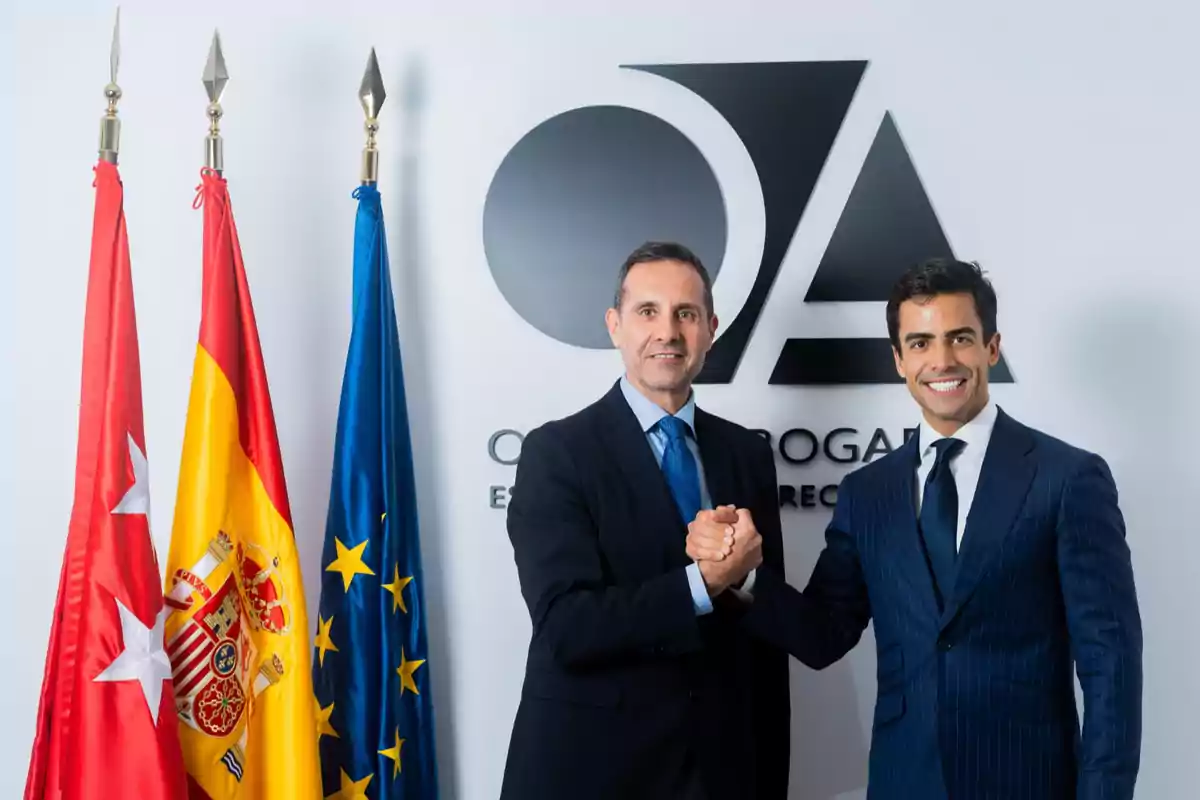 Dos hombres de traje estrechando manos frente a un logotipo y tres banderas.