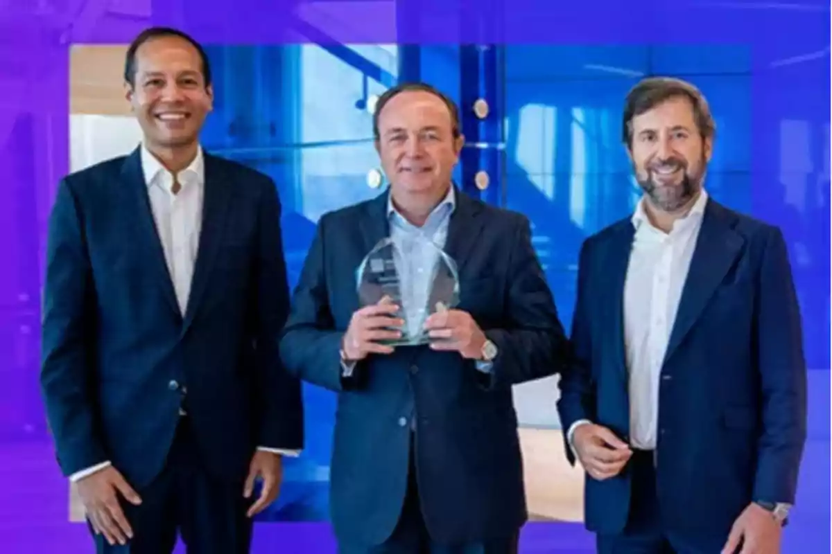 Tres hombres de negocios posan sonrientes, el del centro sostiene un trofeo de cristal, todos visten traje y están frente a un fondo azul Tres hombres de negocios posan sonrientes, el del centro sostiene un trofeo de cristal, todos visten traje y están frente a un fondo azul