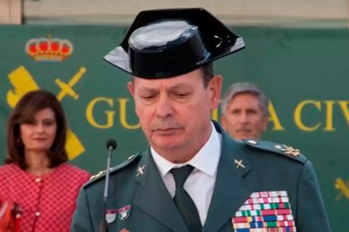 Un oficial de la Guardia Civil con uniforme y medallas, hablando en un micrófono durante un evento oficial, con un fondo verde que incluye el emblema de la Guardia Civil y dos personas en segundo plano.