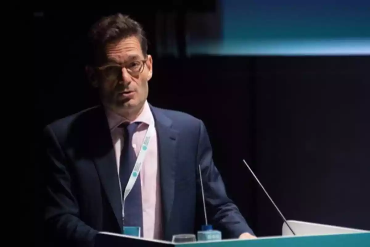 Un hombre con gafas y traje oscuro habla en un podio durante una presentación o conferencia. Un hombre con gafas y traje oscuro habla en un podio durante una presentación o conferencia.