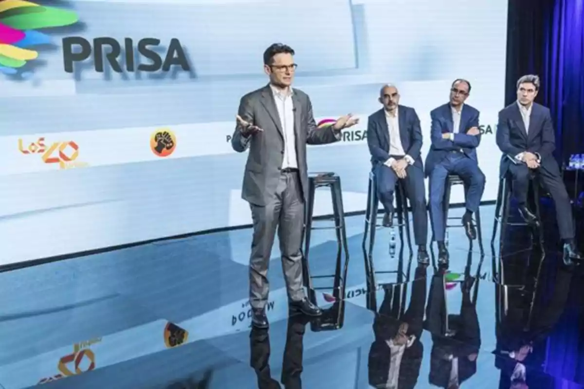 Un grupo de hombres en trajes formales participa en una presentación de PRISA, con logotipos de Los 40 y Cadena Dial visibles en el fondo.