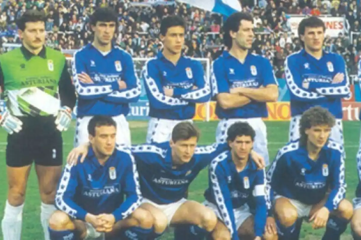 Un equipo de fútbol posando en el campo con uniformes azules y un portero con uniforme verde frente a una multitud de espectadores.