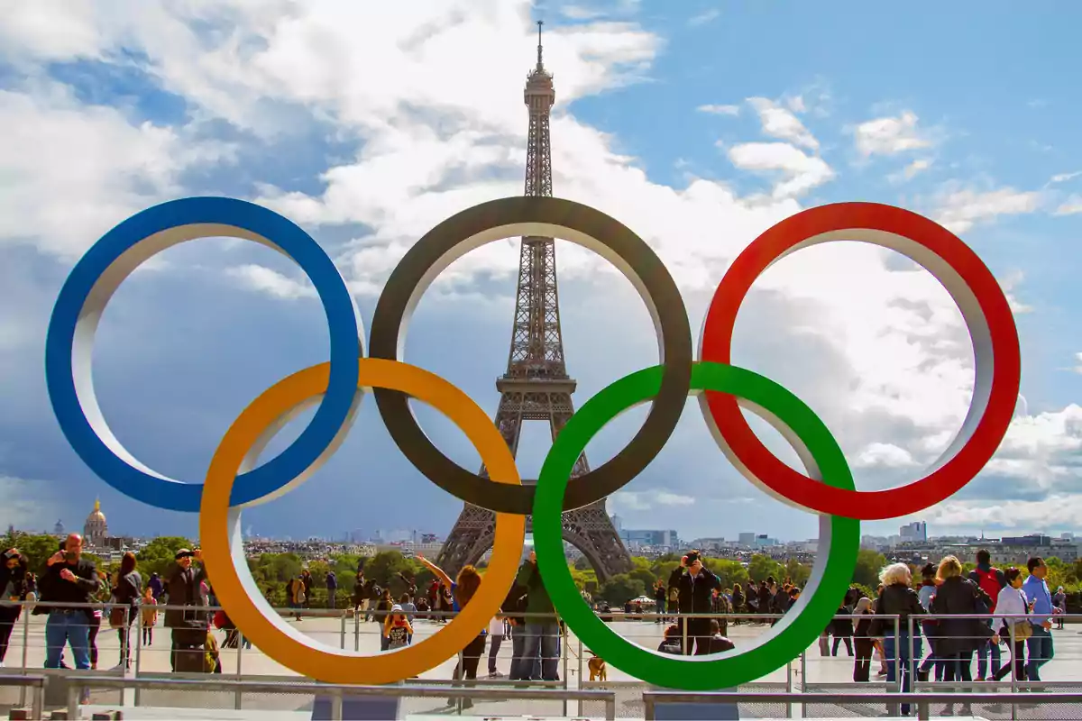 Anillos olímpicos frente a la Torre Eiffel en París con personas alrededor y cielo parcialmente nublado.