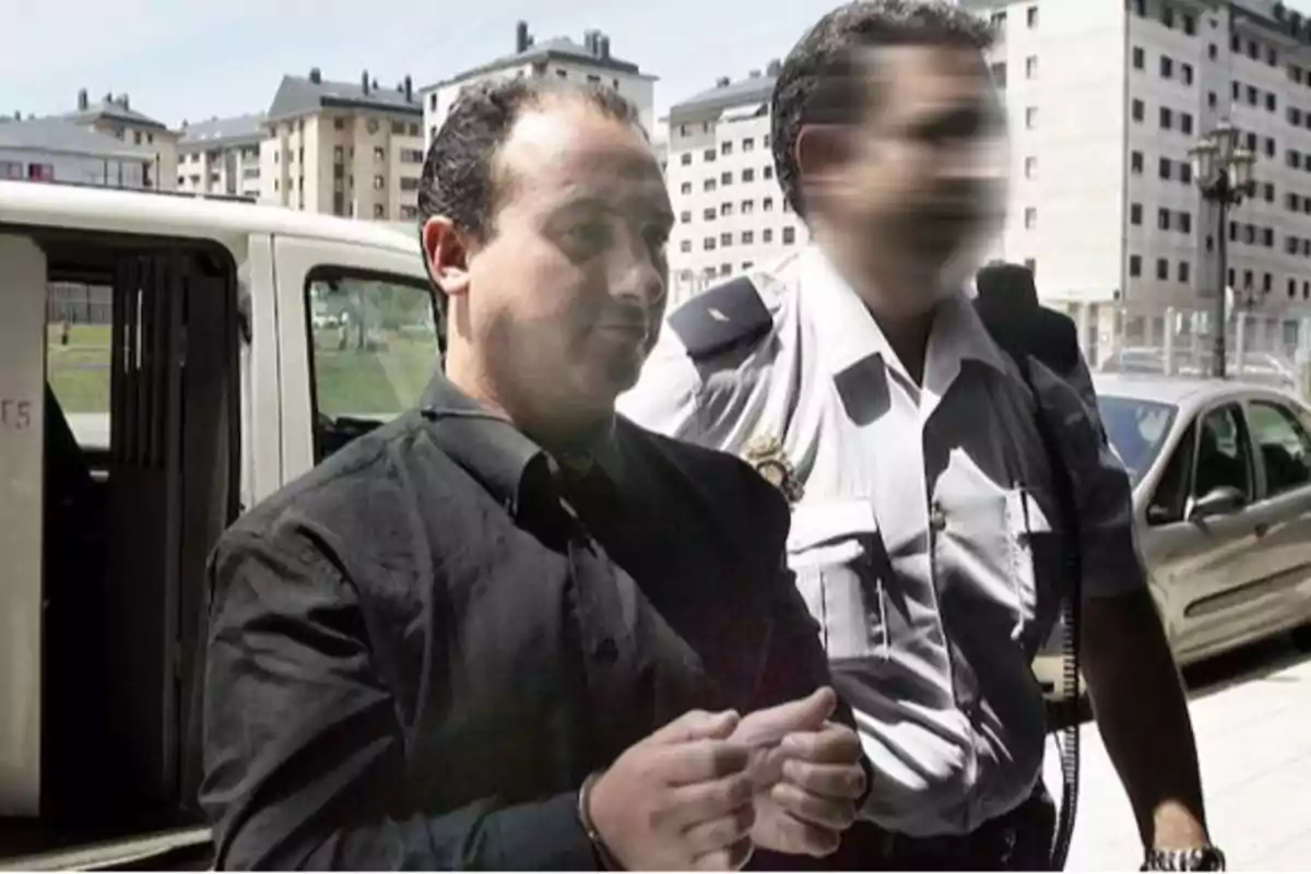 Un hombre esposado es escoltado por un oficial de policía con el rostro difuminado, mientras caminan junto a un vehículo en una zona urbana con edificios de apartamentos al fondo.