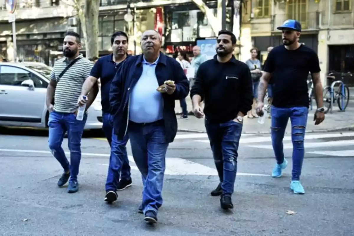 Un grupo de cinco hombres caminando por una calle urbana, con edificios y coches en el fondo.