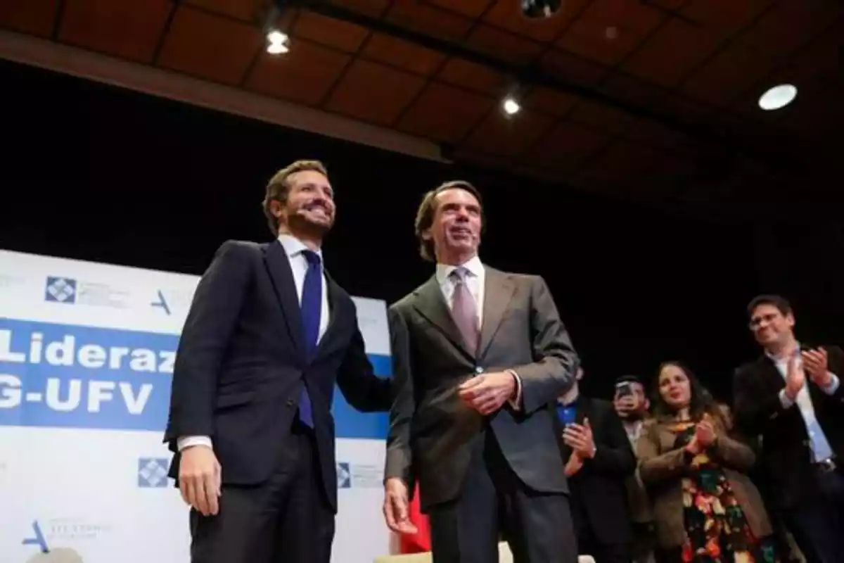 Los exlíderes del Partido Popular, Pablo Casado y José María Aznar en un congreso. En la foto, sacada desde abajo, sonríen e la cámara. Pablo Casado tiene la mano en la espalda de Aznar y ambos visten trajes.