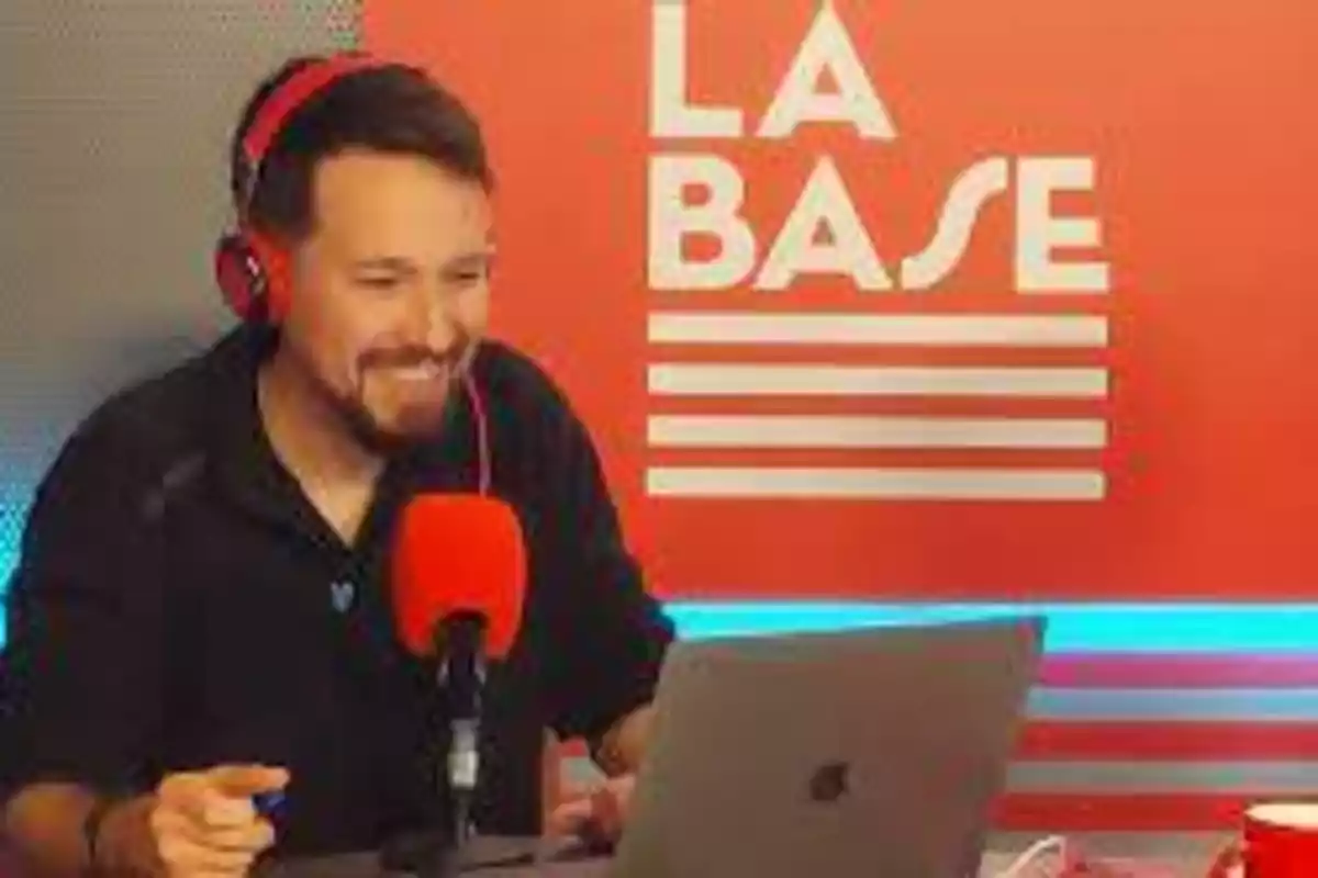 Un hombre con auriculares rojos y un micrófono rojo está sentado frente a una computadora portátil, con un fondo rojo que tiene el texto "LA BASE".